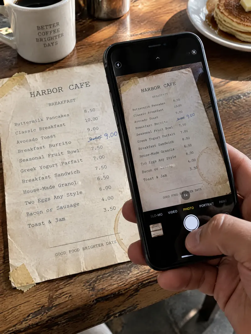 Een hand houdt een telefoon boven een versleten papieren menukaart op een cafétoonbank om er een foto van te maken.