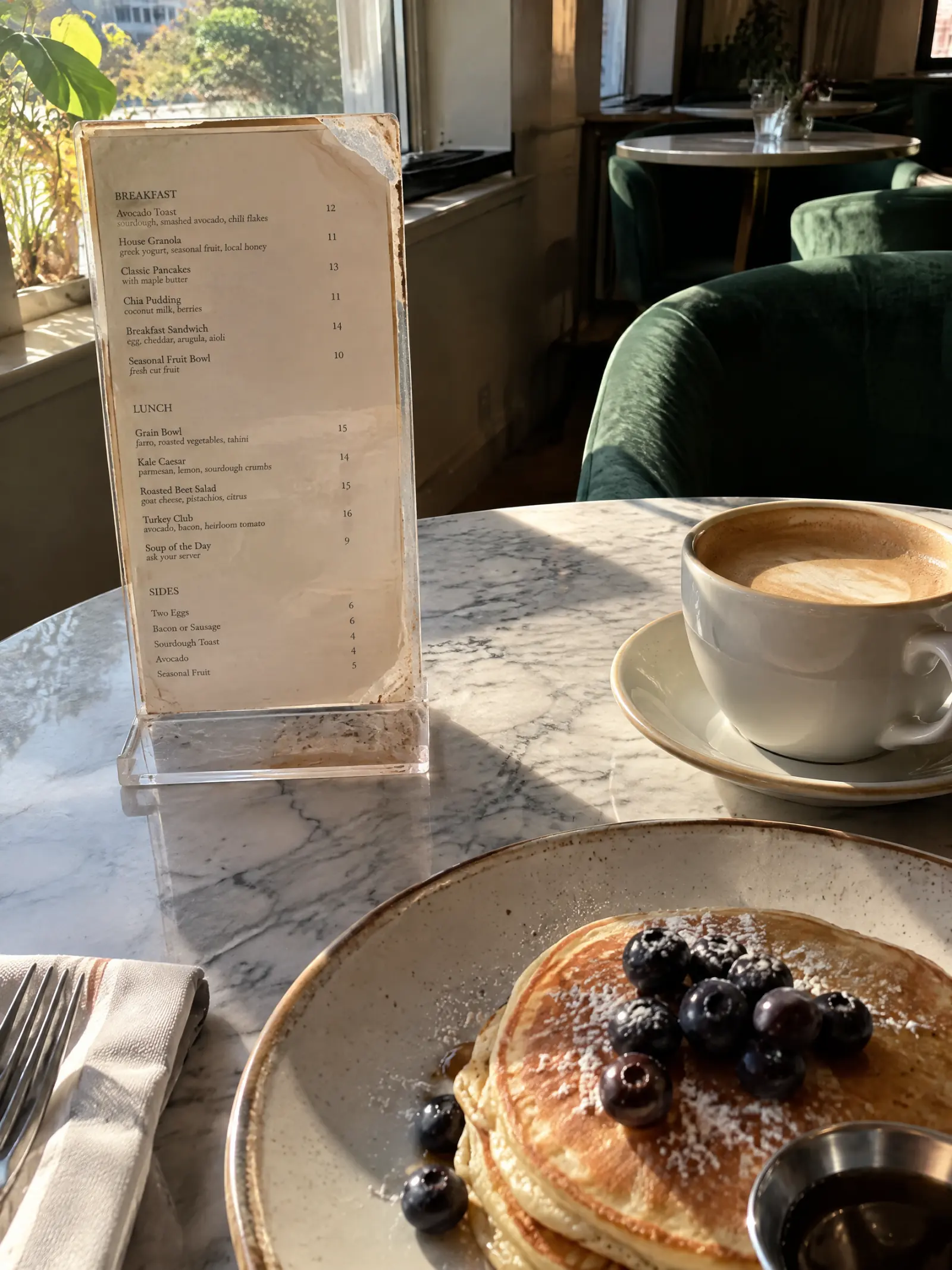 Et cafébord med et gammelt lamineret menukort i en ridset plastikholder, en kop kaffe og en tallerken pandekager.